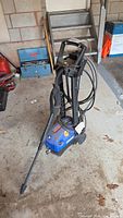 Full view of the blue Simoniz pressure washer unit standing on a concrete garage floor with hose wrapped around handle.