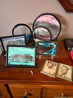 View of six sand art pieces on a wooden surface, including two circular pieces on black swivel stands and four rectangular framed pieces, two small.