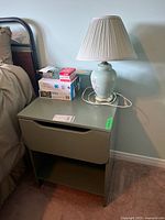 Green veneer side table with drawer and lower shelf next to bed with lamp on top.