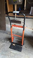 Photo of front angled view of metal dolly with black handle and orange frame, showing foldable base plate and wheels.