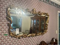 Full view of the vintage ornate gold framed rectangular wall mirror hanging against patterned wallpaper showing reflection of dining room.