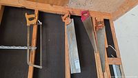 Four vintage hand saws with wood handles hanging on wall frame showing varying sizes and designs.