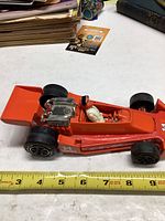 Side view of orange race car with driver, showing length near measuring tape for size reference