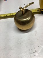Photo of solid brass apple-shaped paperweight on a white surface with a yellow measuring tape showing approximately 3-inch height.