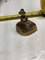 Brass bell figurine shaped like a woman with a wide skirt and hat placed next to a measuring tape showing approx. 3 inches in size.