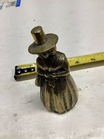 Brass bell shaped like a woman in traditional dress with tall top hat, placed next to a tape measure showing height around 4.5 inches.