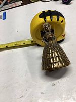 Brass bell shaped like a woman in skirt next to a yellow tape measure showing approximately 5 inches height