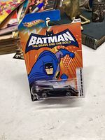 Unopened 2011 Hot Wheels Batman The Brave and the Bold die-cast car in original blister card packaging on white surface with background items visible.