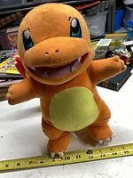 Front view of the orange Charmander plush with cream belly and large embroidered eyes, sitting upright on a flat surface with a measuring tape showing about 10 inches tall.