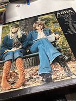 Front cover of the ABBA Greatest Hits vinyl record showing two band members seated on a park bench in autumn, wearing casual fall clothing.