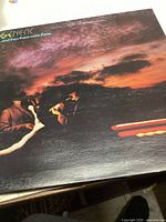 Front cover of Genesis vinyl album '…And Then There Were Three' showing three band members and a dark cloudy sky with light streaks.