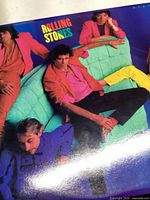 Front cover of the 'Dirty Work' album by The Rolling Stones showing band members sitting on a turquoise couch in colorful clothes.