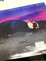Front cover of Stevie Wonder's 'In Square Circle' vinyl record featuring the artist sitting next to a large circular object on a rocky landscape with a purple sky background.