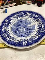 Top view of the round ceramic plate showing blue and white detailed country scene with horse-drawn wagon and floral border