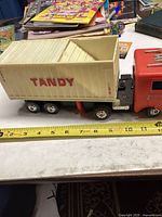 Side view of plastic transport truck showing red cab and cream container with TANDY lettering. Measuring tape indicates length about 11 inches.