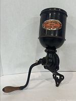 Front-side view of the Universal 014 Coffee Mill Grinder showing the red and white label on the hopper and the curved wooden handle crank attached to the black cast metal grinder body.