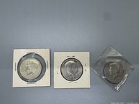 Three worn Kennedy Half Dollars, two in cardboard holders and one in plastic sleeve, showing obverse side with Kennedy profile