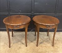 Pair of antique French Provincial wooden side tables with scalloped edges and single drawer with decorative metal handle. Visible wear on the wood surface.