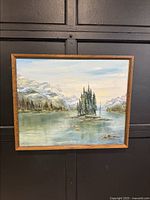 Front view of the framed canvas painting showing a mountain lake with an island of pine trees and snowy peaks under a partly cloudy sky.