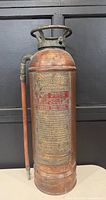 Front view of antique fire extinguisher showing engraved label with red paint detailing usage instructions and manufacturer.