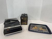Front view of a vintage Panasonic tape recorder, a black box camera with brass front, and Hershey's Kisses metal tray showing advertising artwork of two children sharing candy.