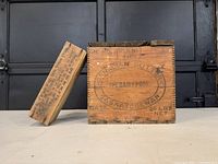 Pair of wooden advertising boxes shown side by side on a flat surface in front of a dark background. The larger box is the Canadian butter box with a lid placed next to it.