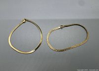 Two gold-tone women's anklets placed flat on a grey surface showing full chain shape and clasps.