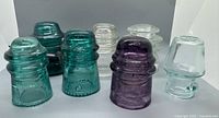 Seven vintage glass telegraph insulators in various colors and designs arranged on a white surface.