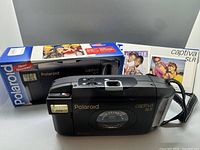 Front view of the black Polaroid Captiva SLR instant film camera with box and manual behind