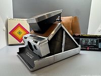 Polaroid SX-70 camera open with leather case, film box, and flash box in background