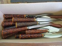 Box with twelve steak knives with faux antler patterned Bakelite handles stacked together showing blade thickness and handle design.