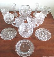 Lot of antique pressed glass children's dishes including handled cups, covered candy dish, candlestick holders, small plates and creamer jug, all clear glass with detailed patterns.