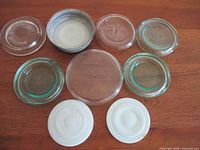 Top view of ten jar lids laid on wooden surface, including nine clear glass lids and one opaque white milk glass lid.