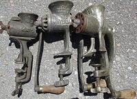 Three antique food grinders shown side by side outdoors on asphalt. Each unit attaches with a clamp and has a hand crank with wooden or metal handles.