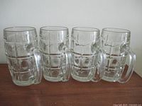 Front view of four clear glass beer mugs with thumbprint pattern and handles, arranged side by side on a white surface.