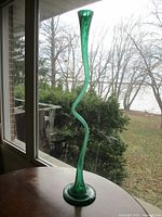 Full height view of tall green Murano blown glass trumpet vase with serpentine curved neck and round base near window with outdoor background