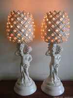 View of two ceramic table lamps lit, showing glowing opalescent bead pineapple shades and cherub bases