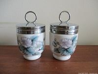 Front view of two Royal Worcester Woodland Flowers egg coddlers side by side on a wooden surface, showing purple floral designs and silver metal lids with loop handles.