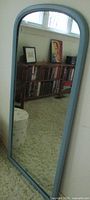 Full view of the vintage full length mirror showing the arched top and Wedgwood blue painted wooden frame, reflecting a bookshelf and artwork in the room.