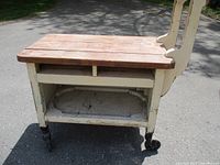 Full side view of vintage wooden trolley cart showing wooden top, open shelf, castor wheels, and painted metal frame with chipped cream paint.