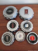 Front view of seven vintage car badge emblems on wooden surface showing logos and condition