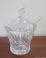 Side view of the lead crystal jam pot with clear intricate cut patterns, bird finial lid, and a crystal spoon inside.
