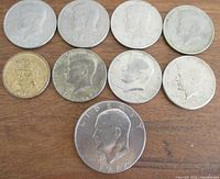 Eight vintage American coins including seven Kennedy half-dollars and one 1972 Eisenhower dollar coin shown on a wooden surface.