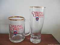 Photo of two Carling O'Keefe beer glasses side by side on flat surface against white wall background. One is a short mug style with gold rim and handle, the other a tall pilsner style with gold rim. Both show red logo and shield on front.