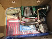 Top-down image inside box showing vintage hand drill, chisels, screwdrivers, measuring tape and other hand tools.