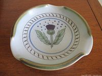 Top view showing round stoneware ashtray with painted thistle and decorative border in blue, green and purple tones.