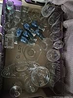 Top view of box showing assorted glassware including shot glasses, blue bottles, pint glasses with print, tumblers, heart dish, platters and stemware