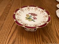 Top view showing floral motif, scalloped burgundy rim with gold accents on pedestal cake plate