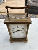 Front view of antique brass carriage clock showing white dial with black Arabic numerals and black hands, brass case, and curved handle on top.
