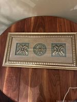 Full view of the framed sculpture showing three metal relief designs on a beige background with gold frame.